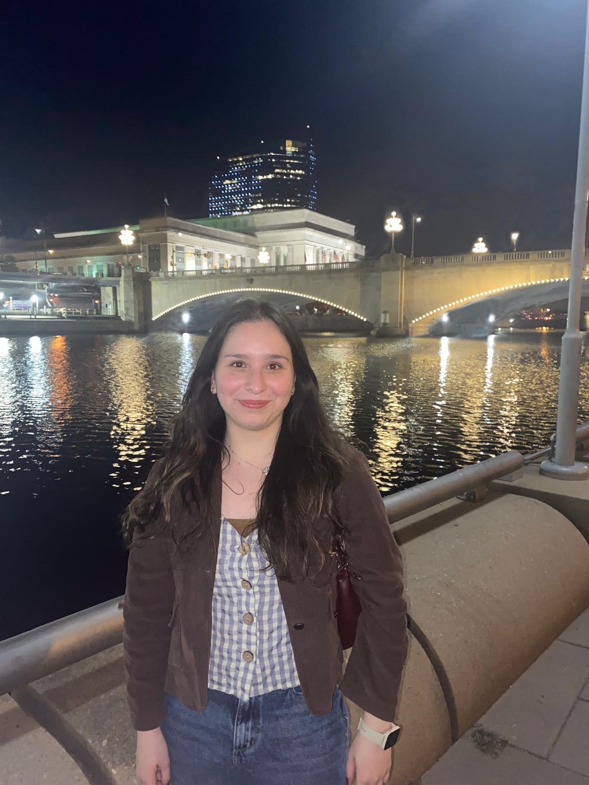 picture of Paule standing outside in a city at night. There's a river and a lit up bridge behind her